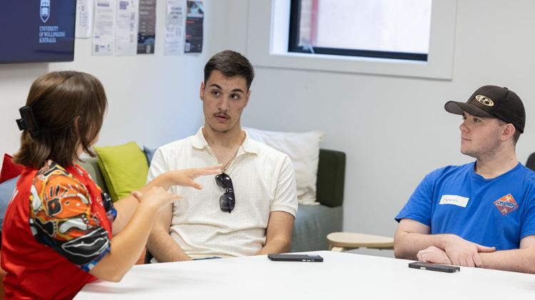 A woman gestures as she speaks to two seated men during a meeting in a modern room with seating and educational posters.
