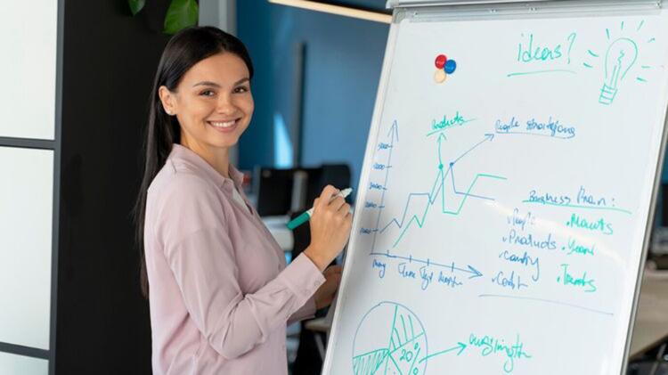 Person smiles while standing next to a flip chart with graphs and notes, holding a marker. Office setting.