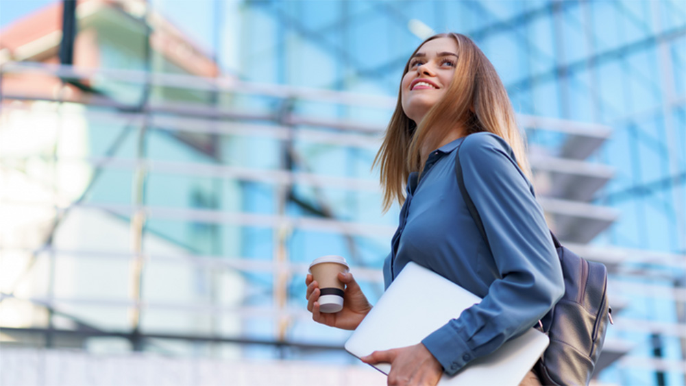 Smiling woman with long hair, in a blue shirt, holds a coffee cup and laptop. She stands outdoors against a modern glass building, exuding confidence.
