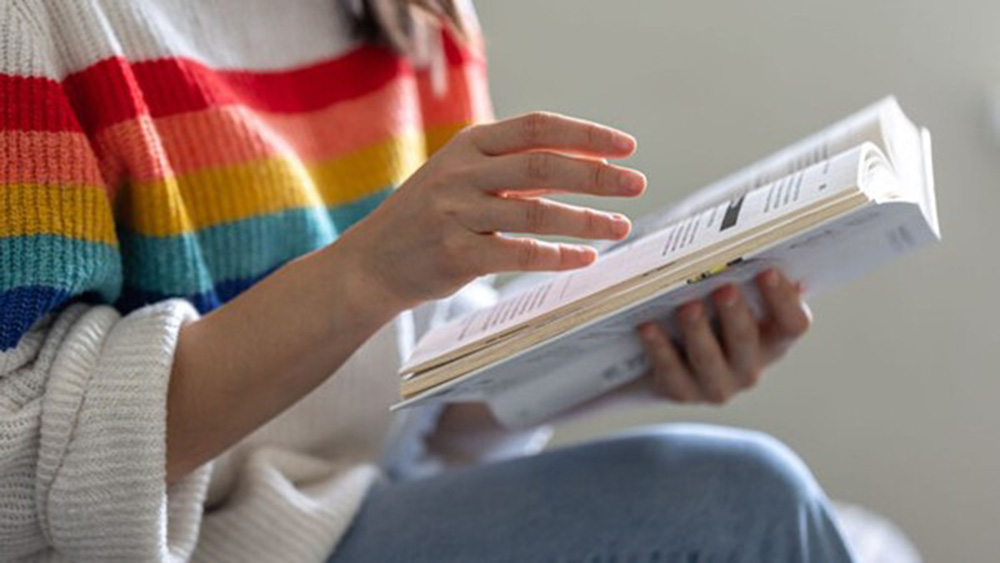 A person in a colorful striped sweater sits, holding an open book and gesturing thoughtfully with their hand.