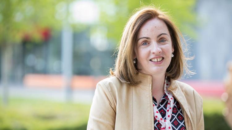 Niamh-Moore-Cherry headshot. Niamh wears a cream suit jacket, a checkered blue and white blouse with flowers in each square, accompanied with a white with red spots neck tie