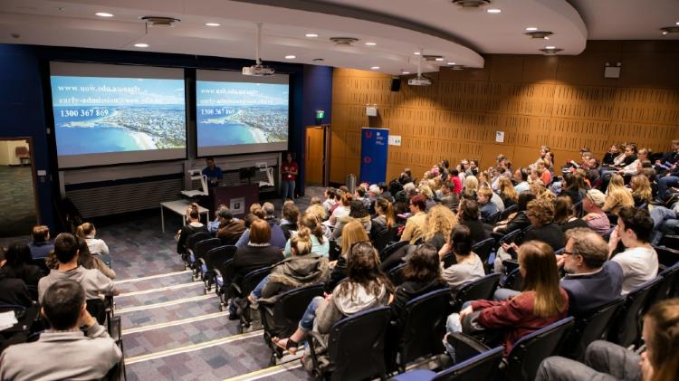 View of a lecture theatre full of people from the back seeing down to the front stage and screen.