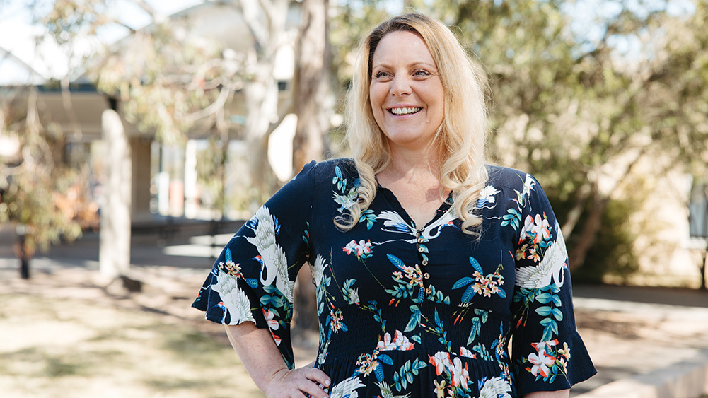 Julia Picton, UOW Master of Education student, standing outdoors on a university campus in a dark floral-patterned dress, with trees and campus buildings blurred in the background.
