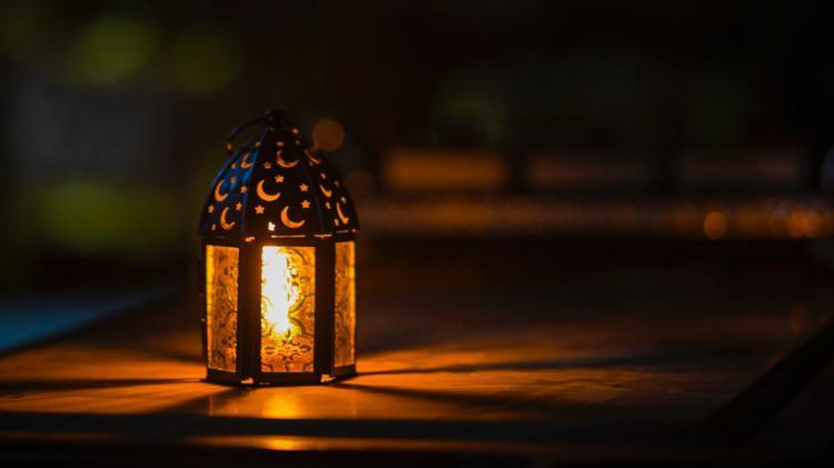 A lantern sits on a table. The lantern in lit up, it has moons and stars cut out of the lid