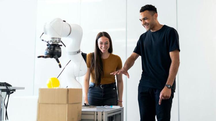 A young woman and a man discuss a robotic arm while interacting with a box and a yellow object in a modern workspace.