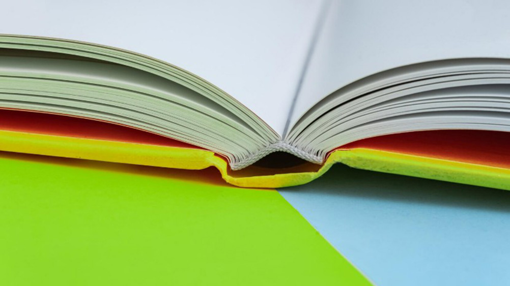 Close-up photograph of an open book viewed from the side, showing curved pages and the book’s spine. Bright yellow and orange endpapers contrast with a green and pale blue background, creating a colourful abstract composition associated with reading, writing and publication.