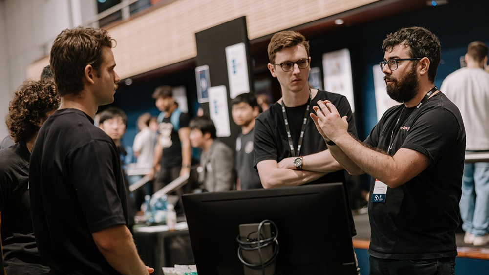 A group of men in black shirts are engaged in a discussion at a tech event. One man gestures expressively, while others listen attentively in a busy, indoor setting.