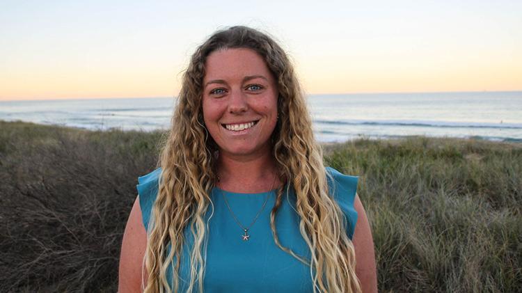 Samara Brown smiles at camera, with the beach in the background. Samara has long, blonde, curly hair and is wearing an aqua coloured tank top