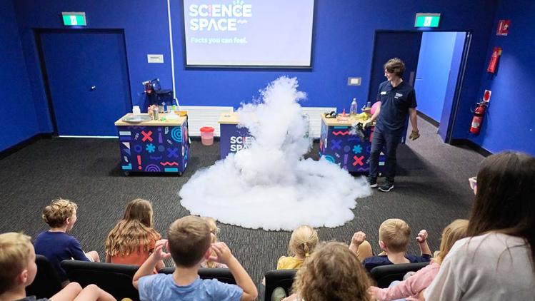A science demonstration in front of an audience of children, featuring a dramatic cloud of smoke or vapor rising from the center of the stage. A staff member in a dark shirt stands to the side, watching the reaction with a smile. The setup includes colorful tables with science equipment and a projection screen displaying the words 'Science Space - Facts you can feel' against a blue wall.