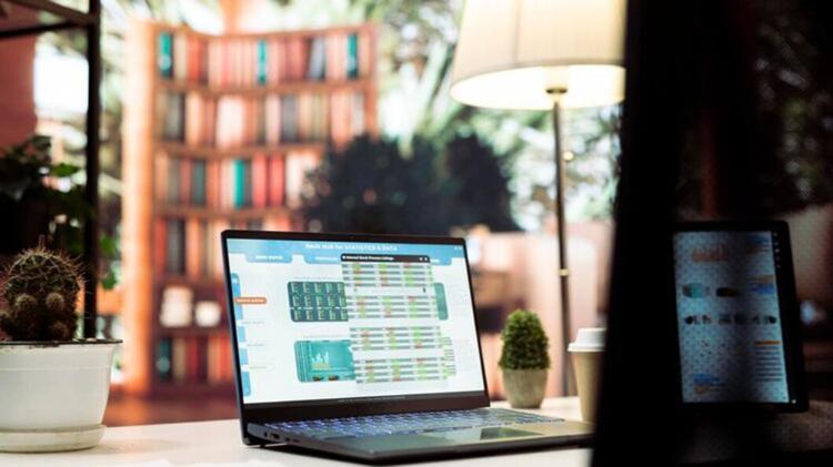 A laptop displays data analytics, with a plant, coffee cup, and bookshelf blurred in the background, creating a cozy workspace atmosphere.