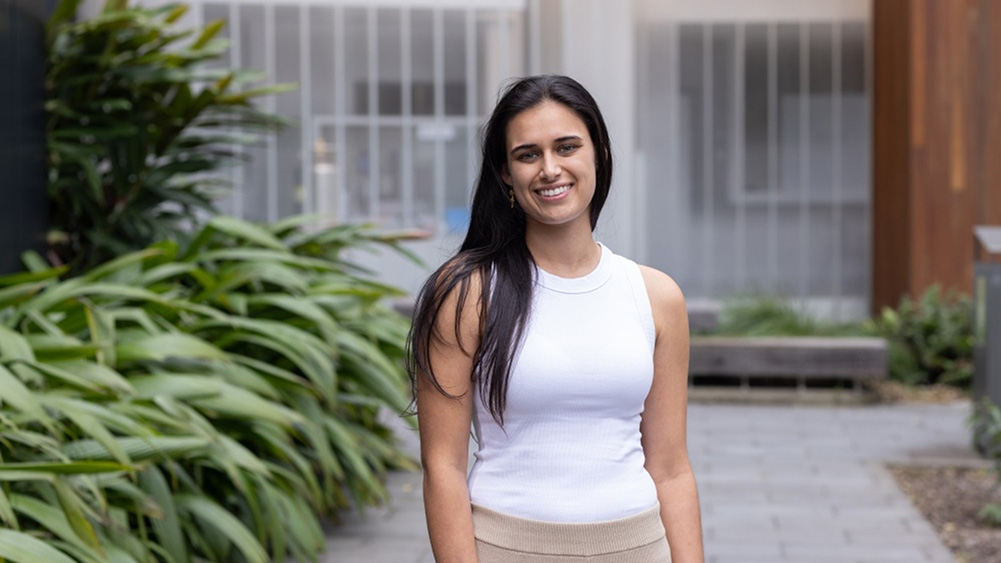 A woman with long dark hair smiles outdoors, wearing a white sleeveless top. She stands on a paved path with lush green plants on the left, conveying a cheerful and relaxed mood.