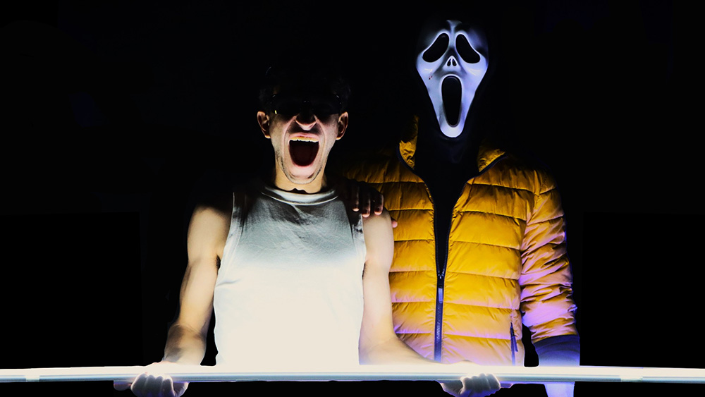 Two people stand side by side in the dark; one wears a white tank top and screams, while the other wears a yellow jacket and a white Ghostface mask.