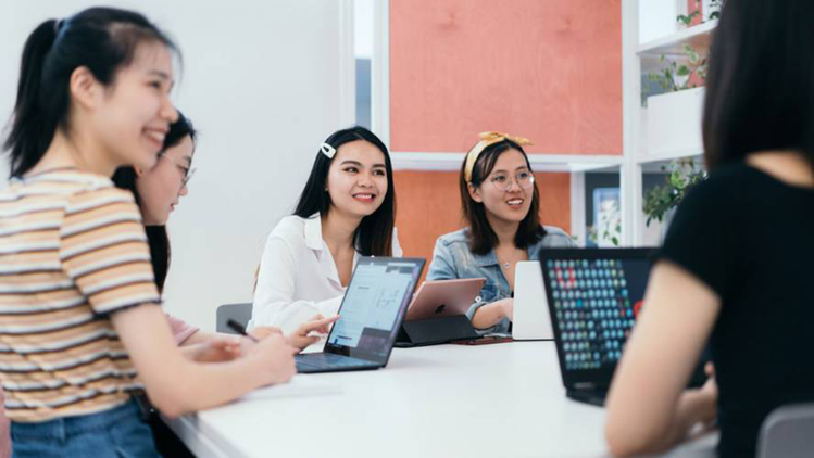 A group of young women sit around a table with laptops and tablets, engaged in discussion and collaboration in a well-lit workspace.