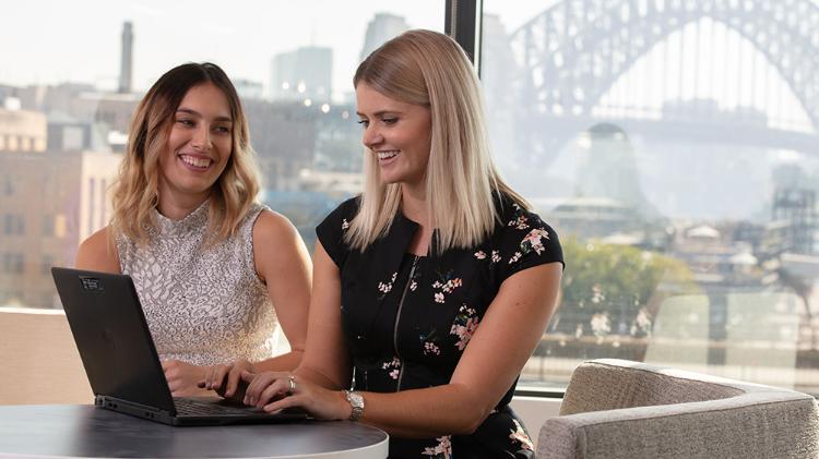 Two people are sitting with laptops smiling as they look at each other.