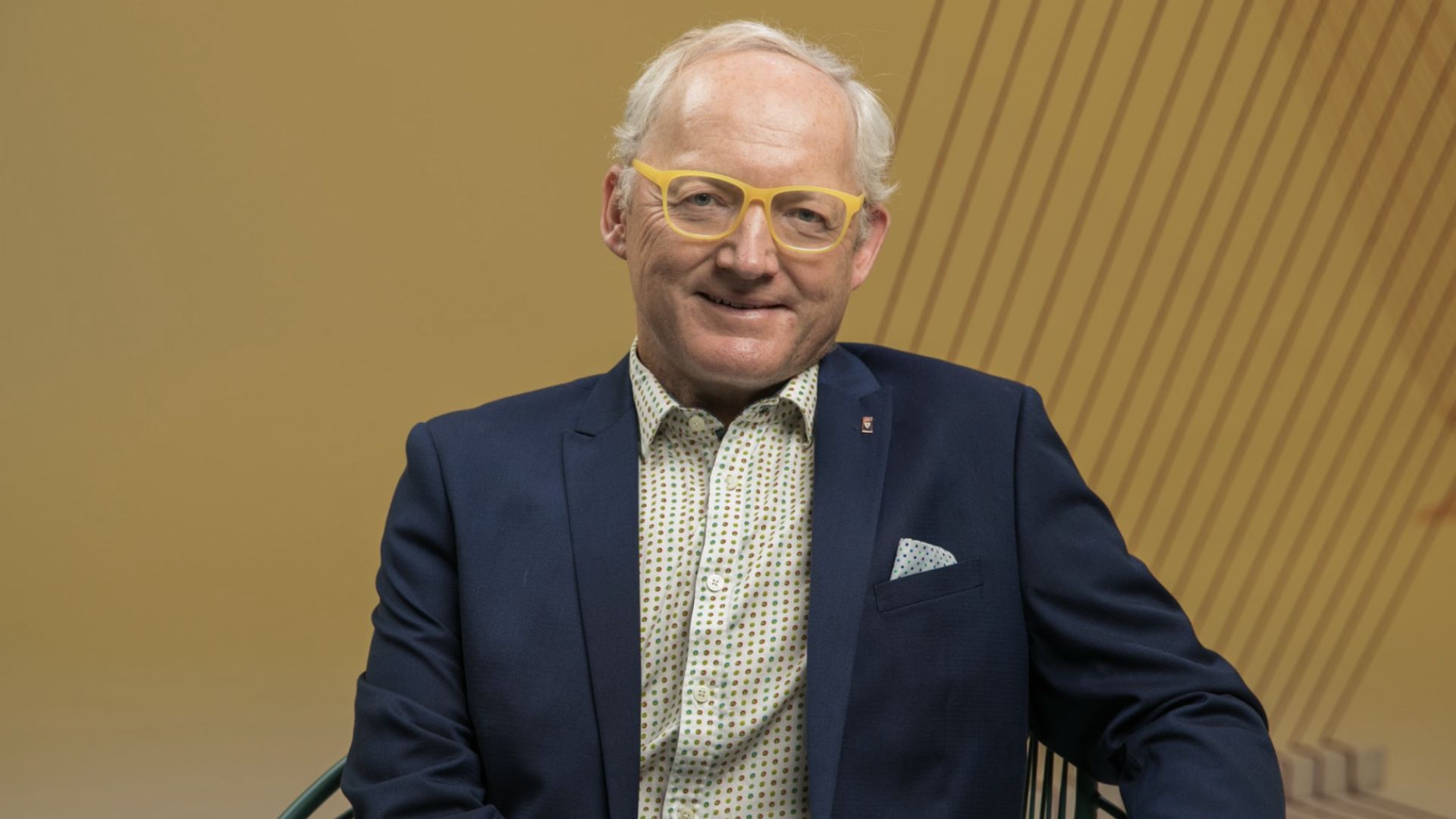 Toby Walsh is sitting in a chair against a mustrad colourd backdrop. He wears a navy suit, with a whit patterned collared button up. His Yellow glasses are a sign of his uplifting personality