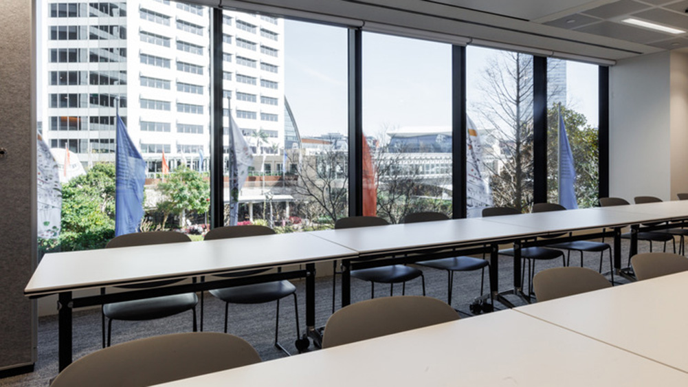 Meeting room with long tables and chairs against large windows. Cityscape and trees visible outside, creating a bright, open atmosphere.