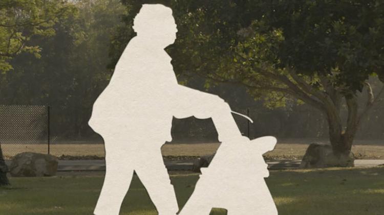 A white cutout a person pushing a walking frame. The background is an outside area with green green and tree bushes.