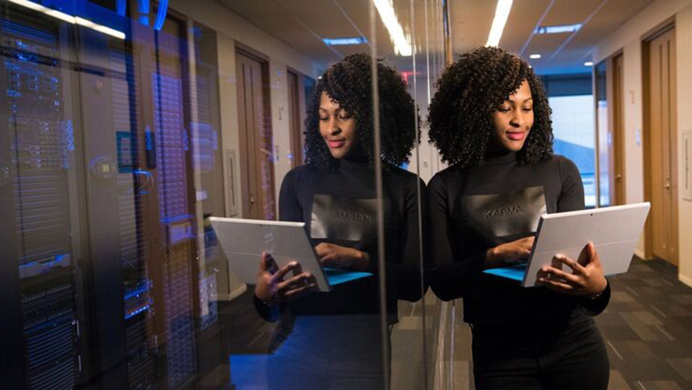 A woman with curly hair holds a tablet in a sleek, modern hallway. She stands next to a reflective glass wall, conveying focus and professionalism.