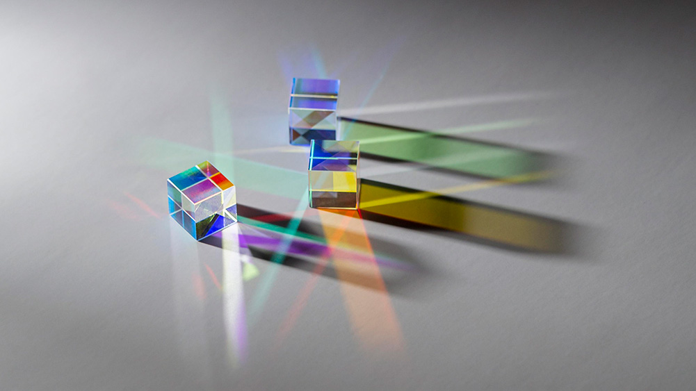 Three transparent glass prisms arranged on a light surface, refracting and dispersing light into vivid rainbow colours. Multicoloured beams and geometric reflections extend across the surface, creating overlapping patterns of red, blue, green, yellow and violet light against a soft grey background. The image illustrates the interaction of light, optics and colour dispersion through prisms.