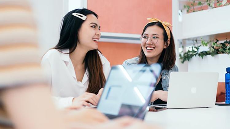 Two people are sitting with laptops smiling as they look at each other.