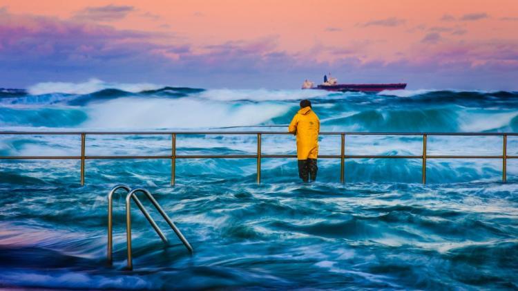 Person in yellow wet jacket standing near the shoreline with rough waves and a cargo ship at Dusk