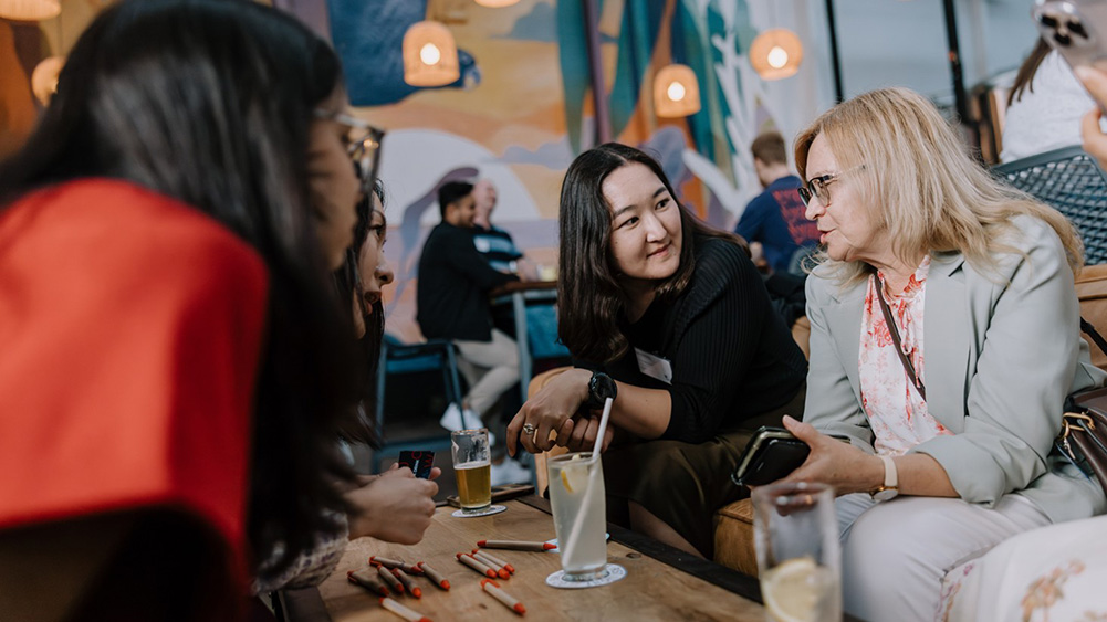 A group of people sit around a wooden table with drinks, engaged in lively conversation. The setting has warm lighting, creating a cozy, relaxed atmosphere.