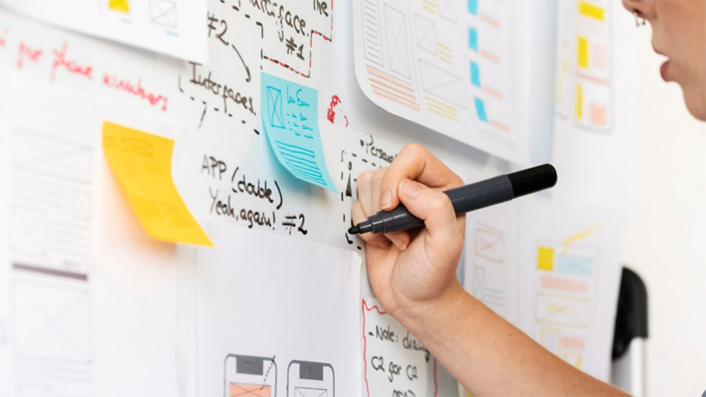 A person writes notes on a whiteboard with colorful sticky notes and wireframe sketches. The scene conveys creativity and collaborative work.