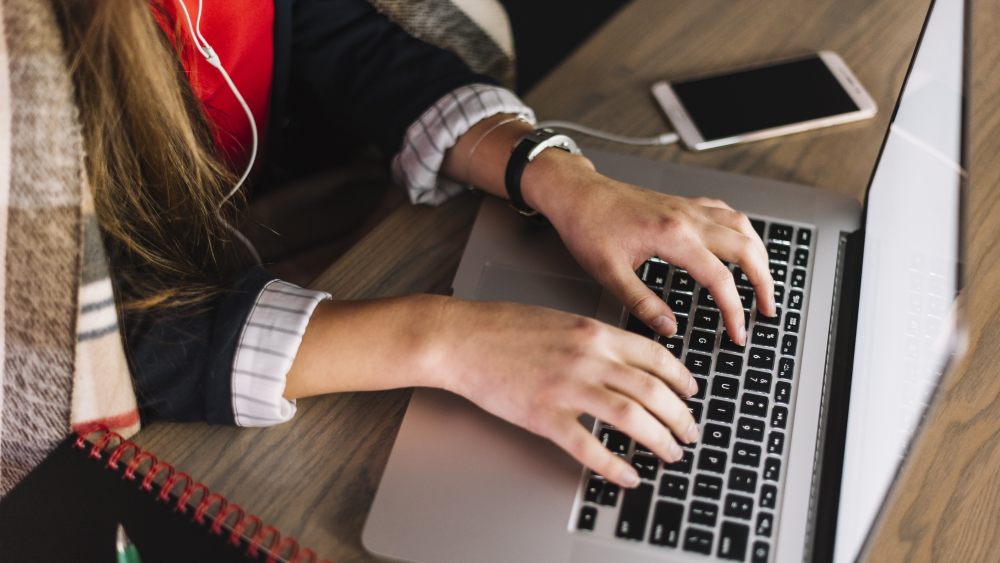 A close-up of a person's hands typing on a laptop, with a smartphone nearby and a cozy scarf draped over the armrest.