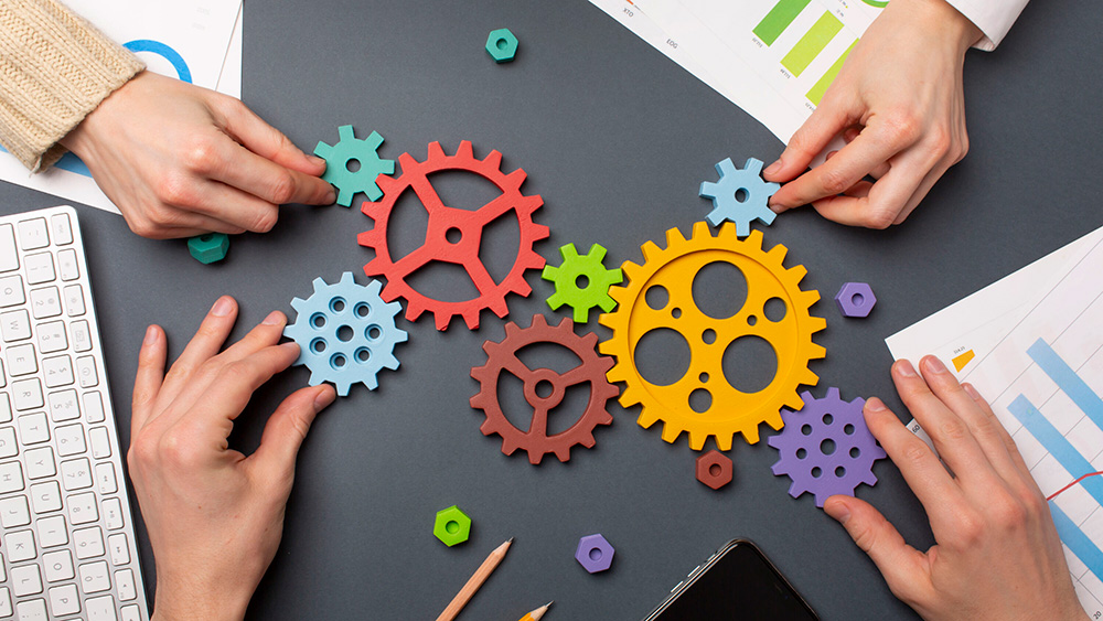 Overhead view of several hands arranging colourful interlocking cogwheels on a desk. Charts, graphs, a keyboard, a smartphone and stationery surround the gears, illustrating project planning, teamwork, collaboration and process management.