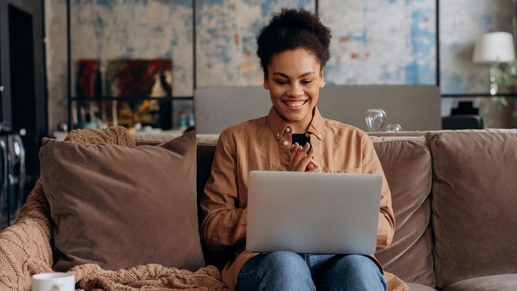 A female smiling and sitting on a couch with a laptop on her knees