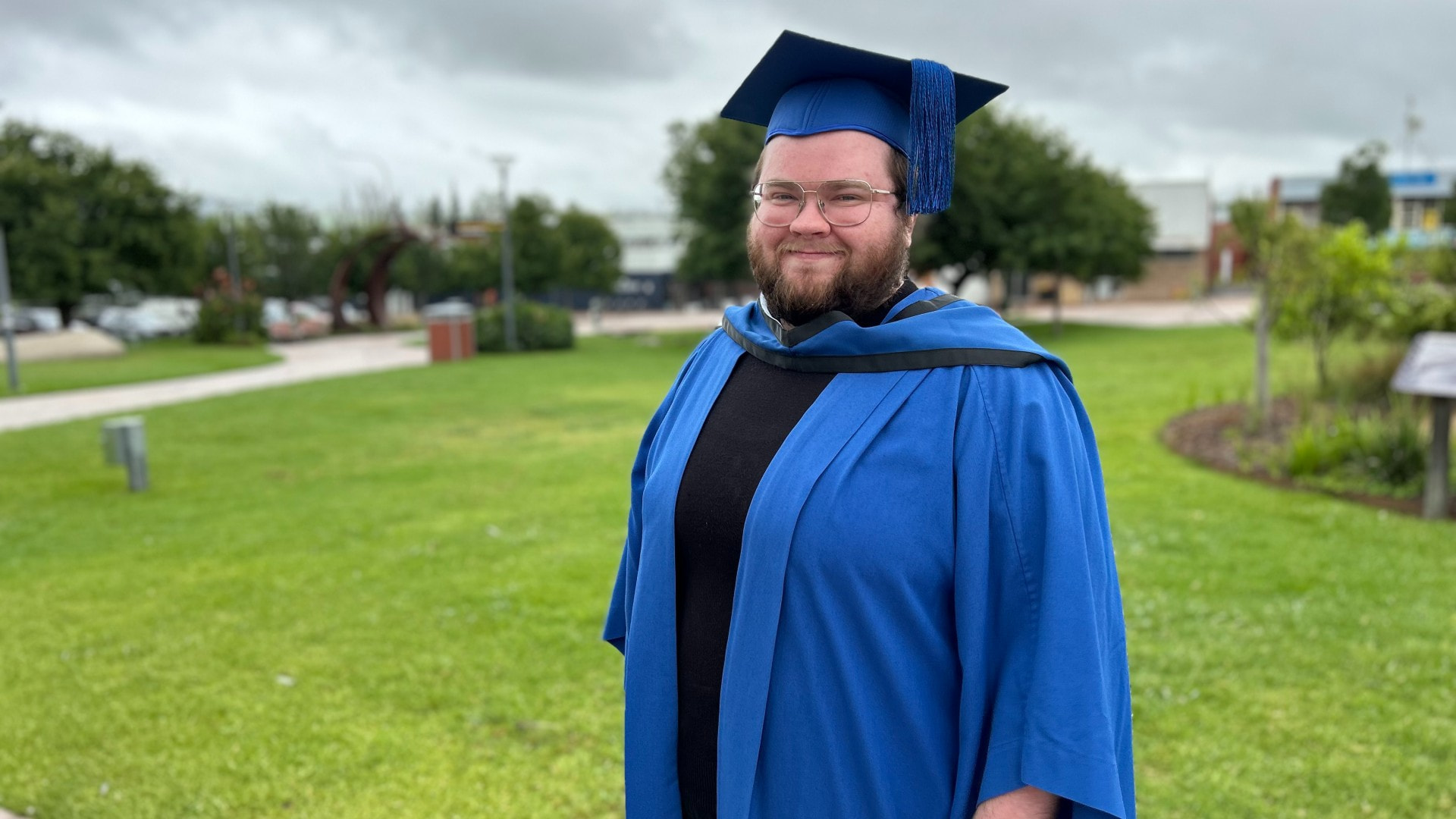 UOW Graduate Adrian Pittman stands in a blue graduation gown and cap. He wears glasses and smiles at the camera.
