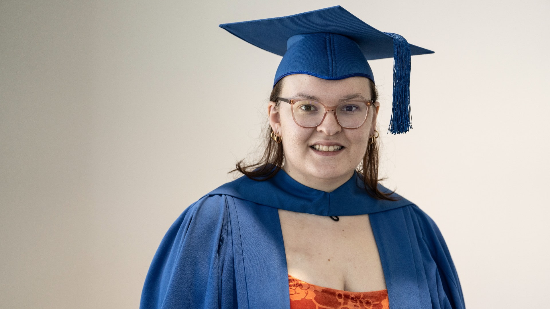 UOW graduate Catie Brown wears a blue graduation cap and stands against a white wall. She is smiling at the camera. Photo: Paul Jones