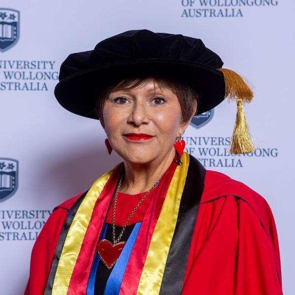 Professor Bronwyn Fredericks wears a black graduation cap and red gown with Indigenous colours in front of a UOW media wall. Photo: Andy Zakeli