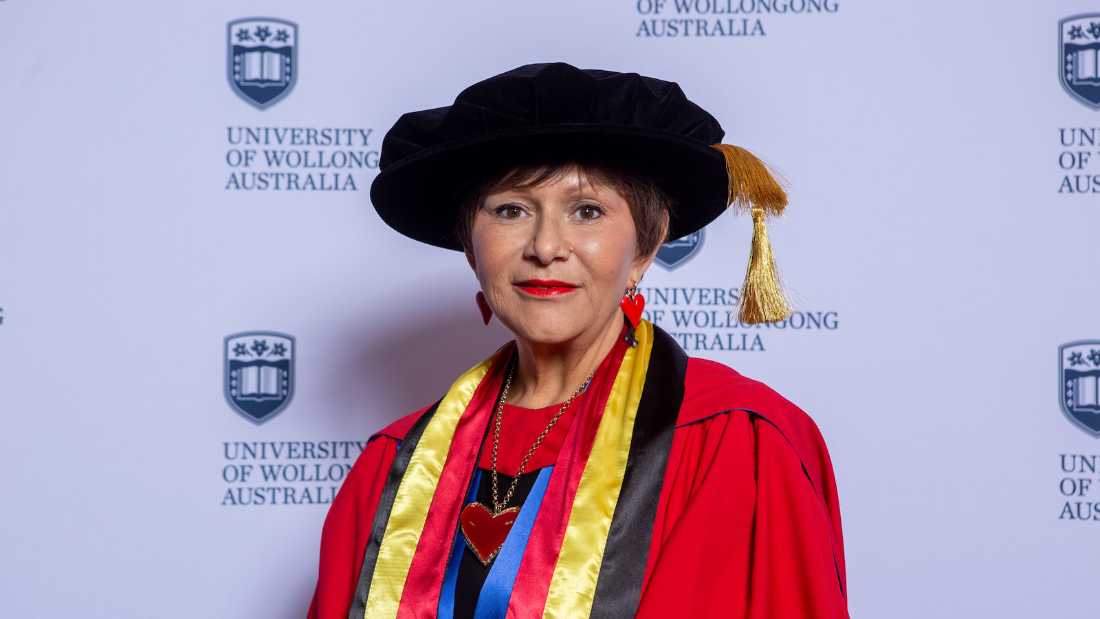 Professor Bronwyn Fredericks wears a black graduation cap and red gown with Indigenous colours in front of a UOW media wall. Photo: Andy Zakeli