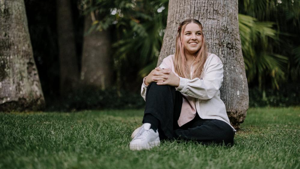 Carly Lavings sits against a palm tree. She wears a light pink jacket and smiles at the camera. Photo: Michael Gray