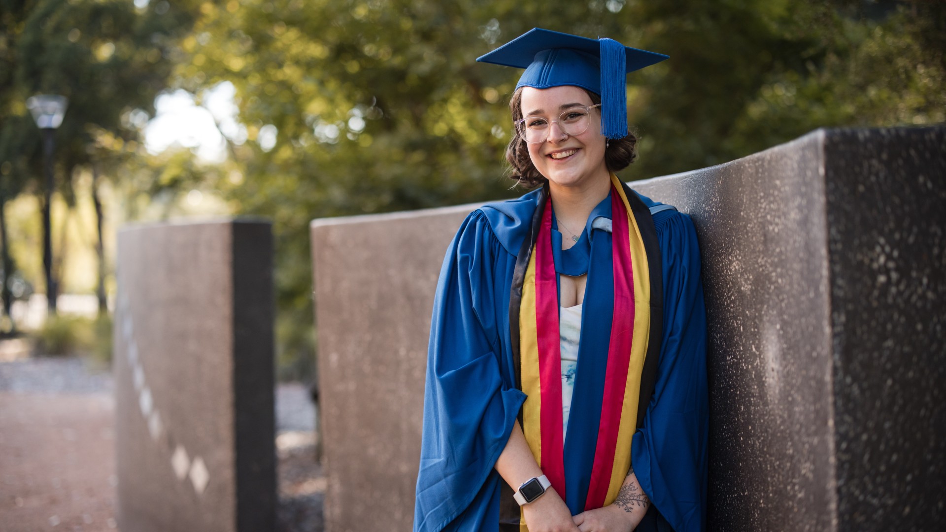 Caitlin Liddle, in a graduation gown with the Indigenous ribbon drapped around her neck, leans against a wall. Photo: Michael Gray