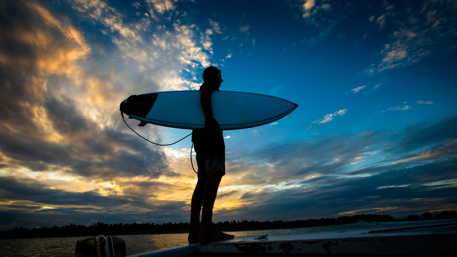 A lone surfer holding a board stands against a sunsetting in the background.