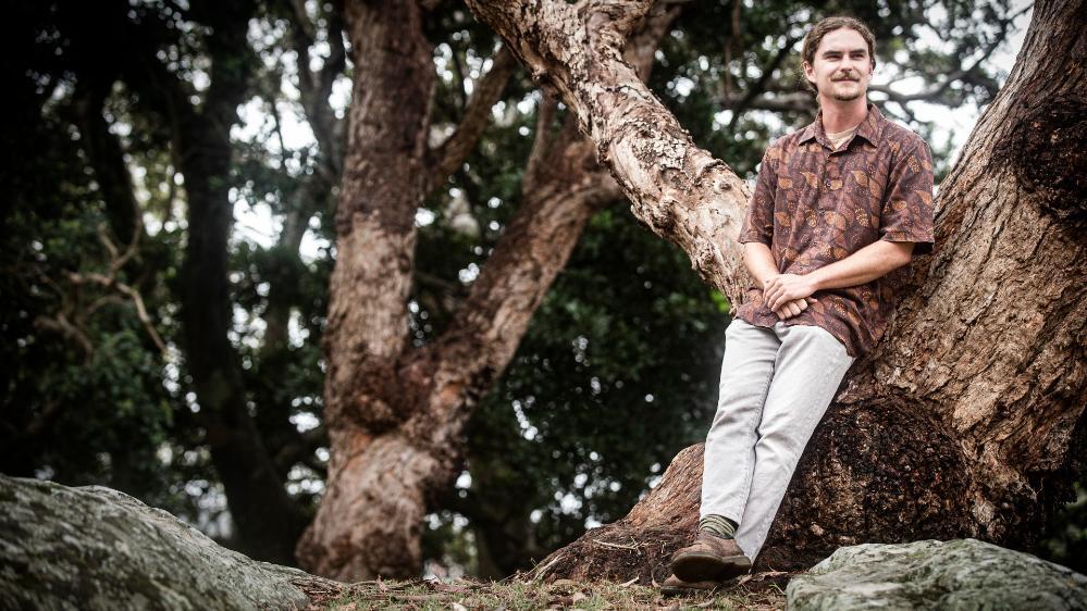 UOW student Charlie Gluskie sits in a tree, surrounded by bush. Photo: Paul Jones