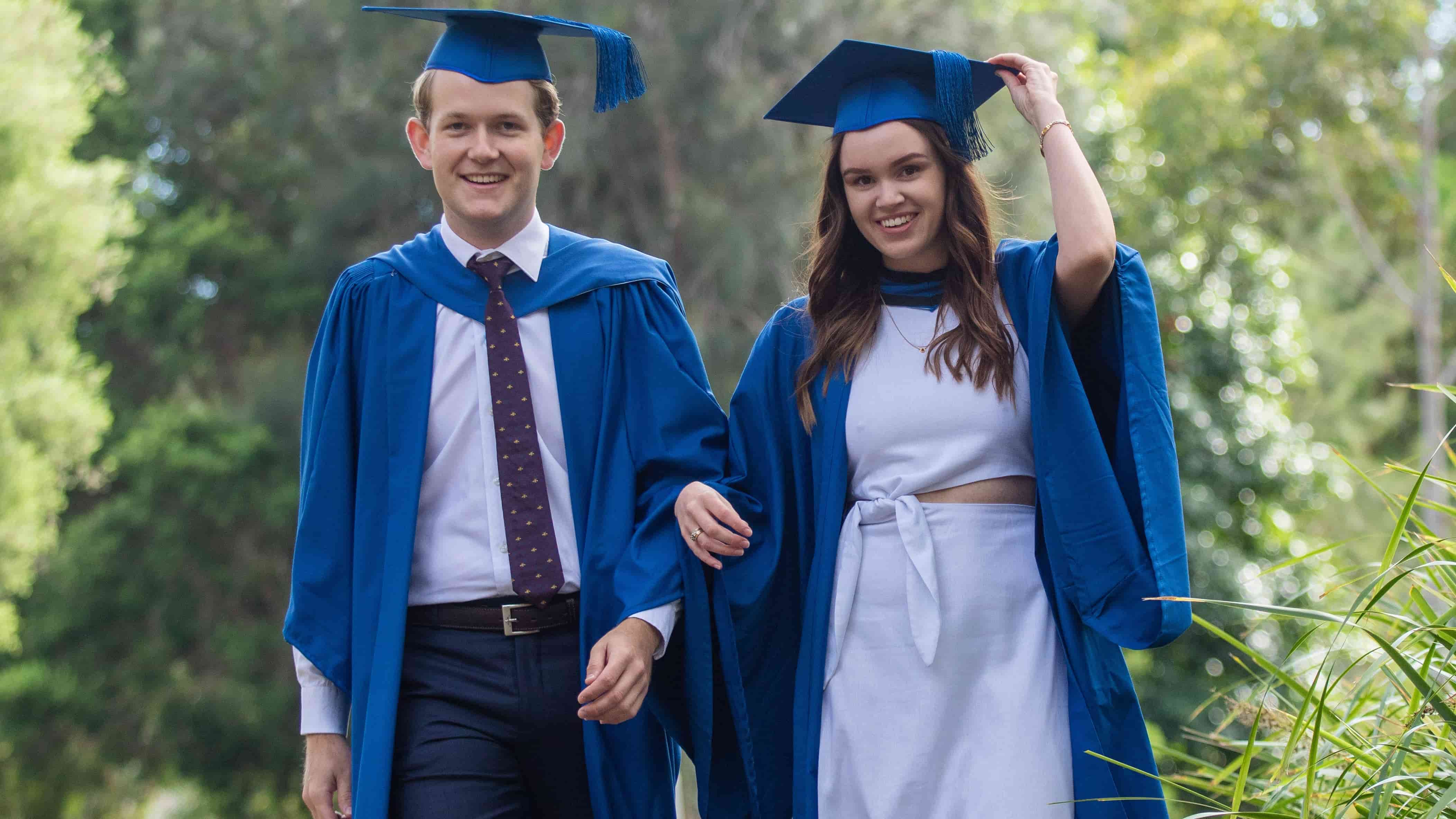 Ruby Evans and Bodhi Morgan both wear blue gowns and mortar boards. They are walking and smiling with linked arms. Photo: Andy Zakeli