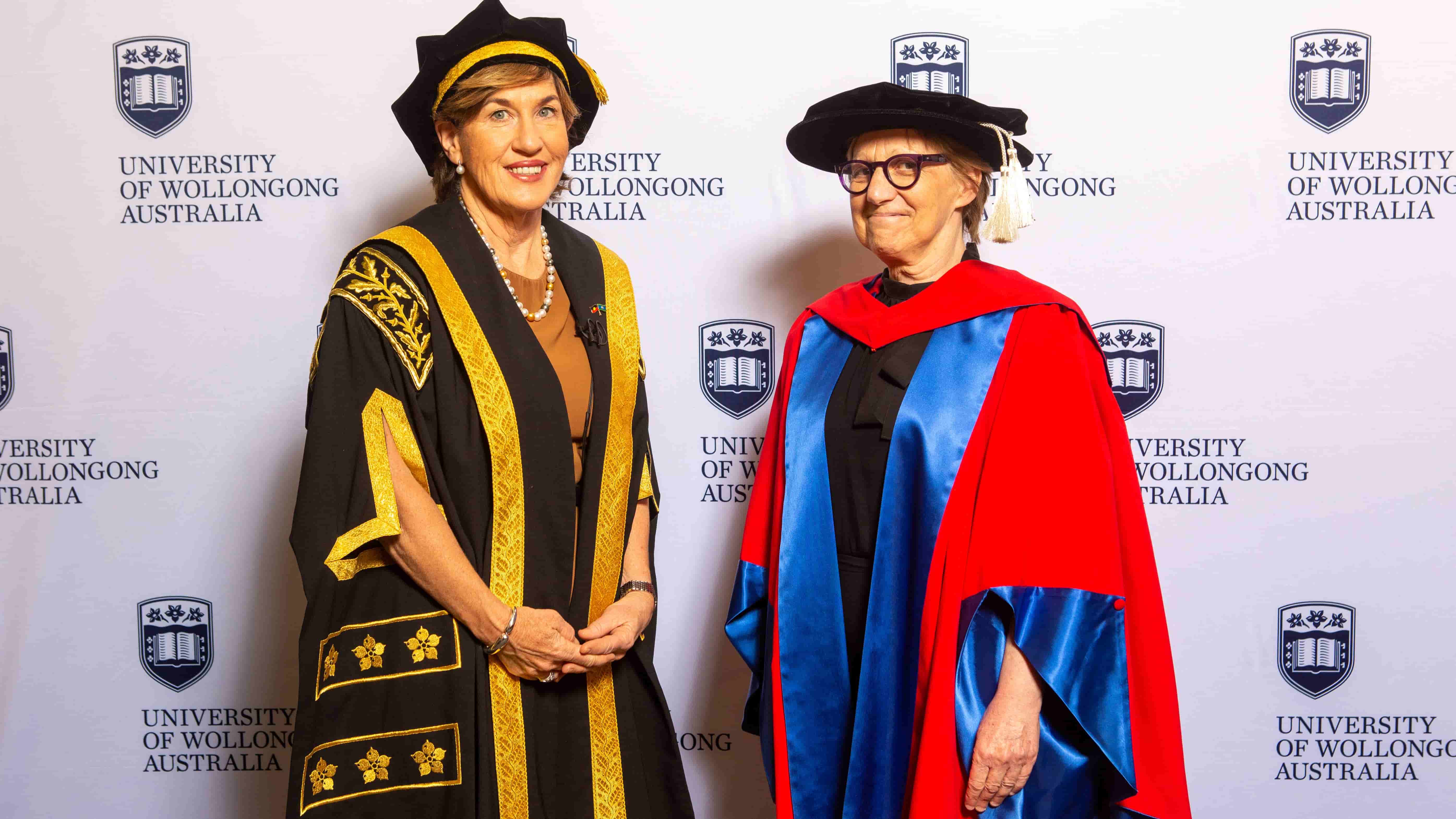 UOW Chancellor Christine McLoughlin and Professor Judy Raper wear graduation gowns and stand in front of a UOW wall. Photo: Andy Zakeli