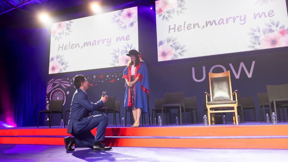 Jin Zhao is on one knee proposing to Helen Qiu, who stands on the graduation stage. On the stage is a slide saying 'Helen, marry me'. Photo: Mark Newsham