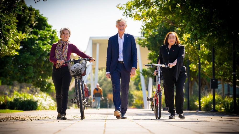 UOW student Juliana Peloche and UOW Vice-Chancellor Professor Patricia Davidson walk either side of Dean Dalla Valle as the trio walk along a footpath at Innovation Campus. Both women are pushing bicycles. Dean does not have a bicycle. Photo: Paul Jones
