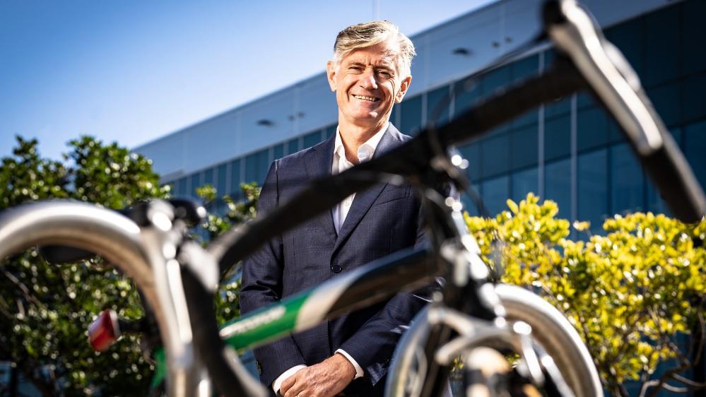 UOW alumni Dean Dalla Valle stands in front of a building on Innovation Campus. In the foreground is the frame of a bicycle. Dean has a smile on his face. Photo: Paul Jones