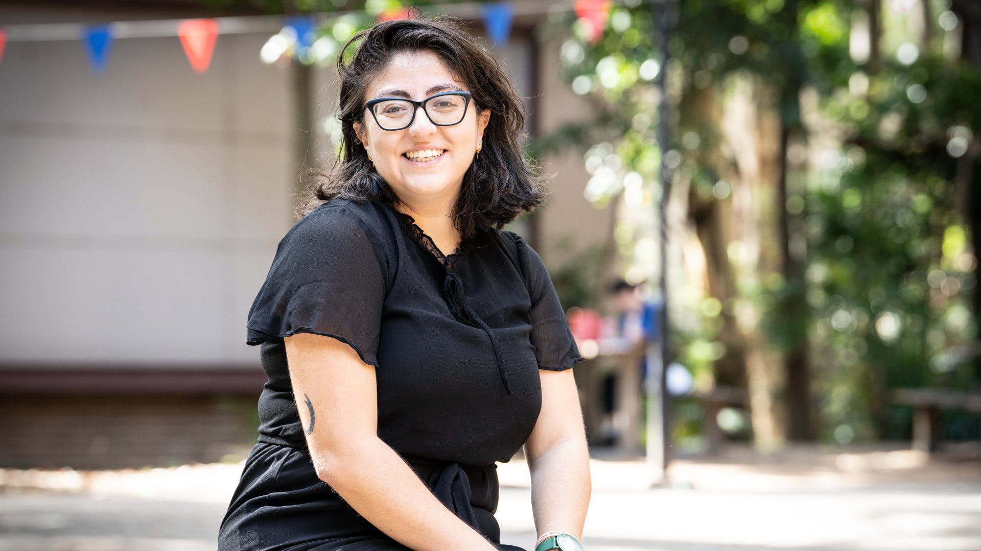Eda Gunaydin wears a black dress and black glasses, sits on a bench and smiles at the camera. The background is slightly blurry. Photo: Paul Jones