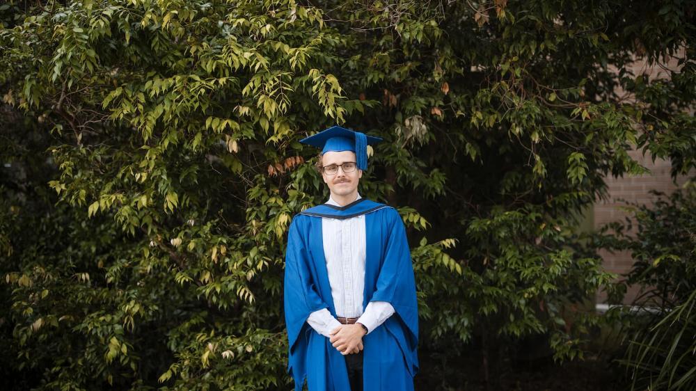 Kiarn Roughley smiles and is wearing a blue graduation cap. He is standing in front of a green bush on Wollongong campus. Photo: Michael Gray