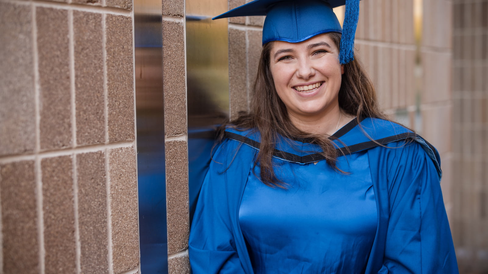 Emma Newman in a blue graduation gown in a close up image. She is leaning up against a wall. Photo: Michael Gray