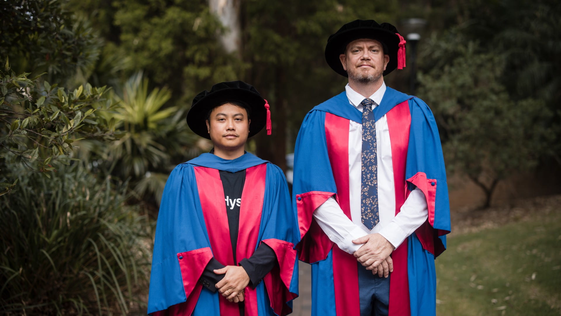 Dr Anh Linh Hoang and Dr Aaron Hodges stand with their hands crossed each in a red and blue graduation gown and cap. Photo: Michael Gray