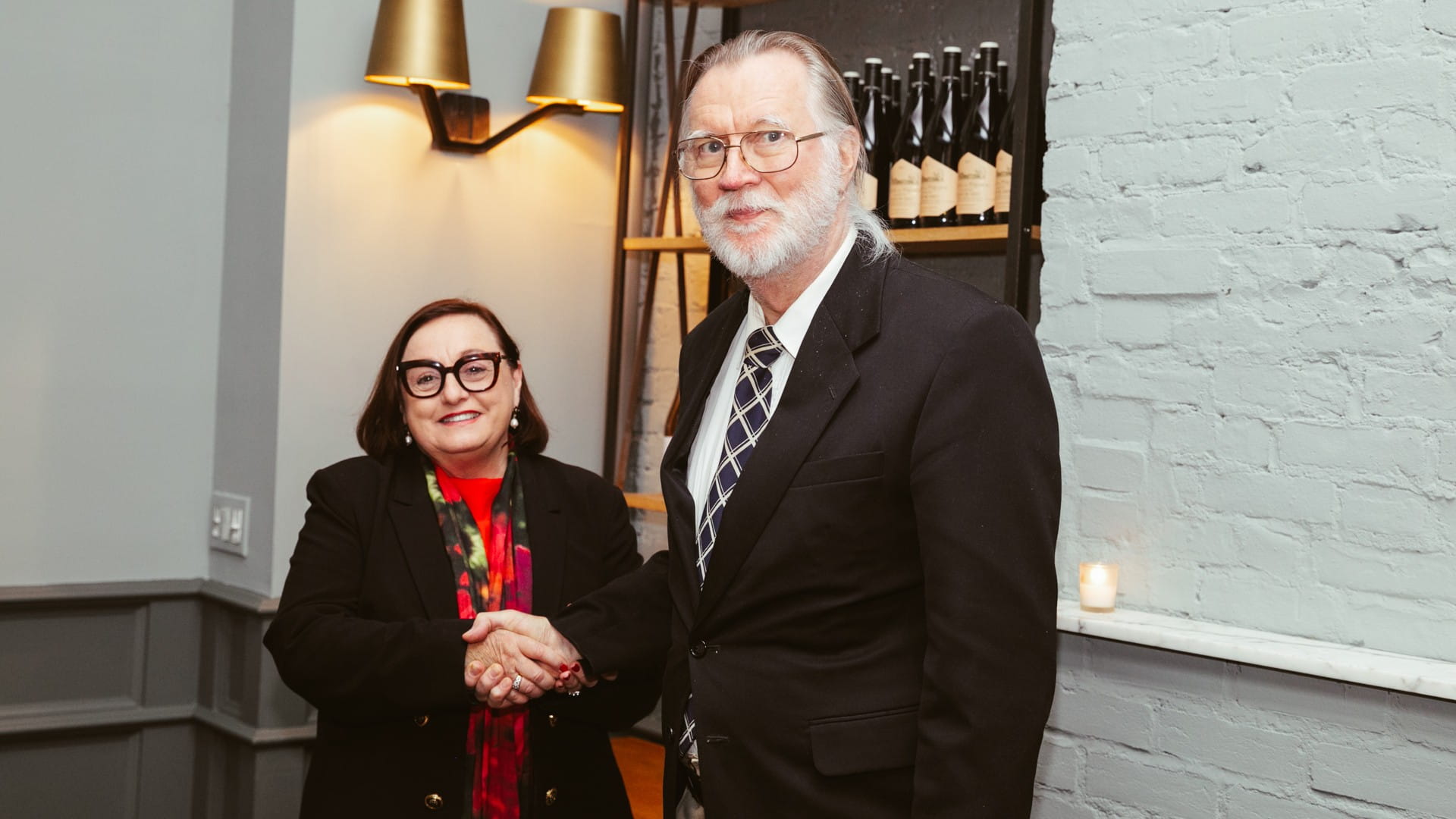 Dr George Delic and Professor Patricia Davidson shake hands and look at the camera. Professor Davidson is to the left and wears glasses and a bright red shirt. Dr Delic wears a dark suit. They are both smiling.