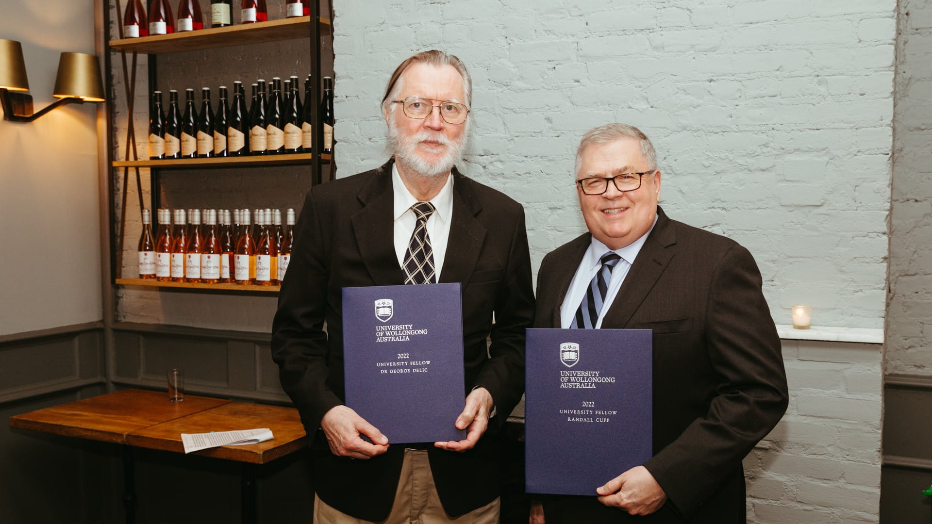 Dr George Delic and Mr Randall Cupp both hold blue books signifying their UOW Fellowships. They stand side by side in a restaurant, against a white wall.