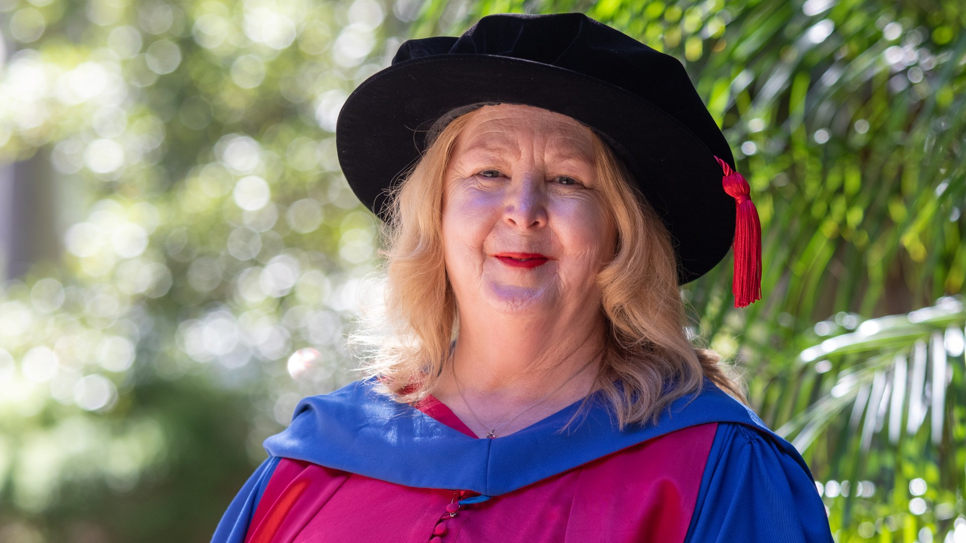 Dr Geraldine Hardie, wearing a blue graduation gown and a black hat, stands in front of a greenery background on graduation day. Photo: Andy Zakeli